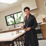Man wearing Jasmisilk men's black pure silk robe with white piping, preparing coffee in a modern kitchen