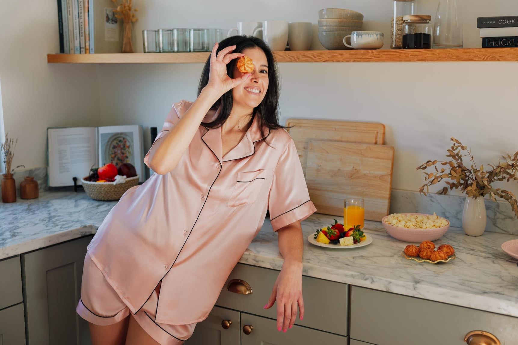 Woman in Jasmisilk silk pajamas enjoying a cookie in the kitchen, combining comfort and everyday luxury.