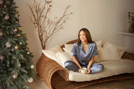 Smiling woman in a luxurious silk pajama set holding a coffee cup while sitting on a sofa next to a beautifully decorated Christmas tree. A cozy holiday moment showcasing the warmth, comfort, and elegance of silk pajamas—perfect for festive gifting.