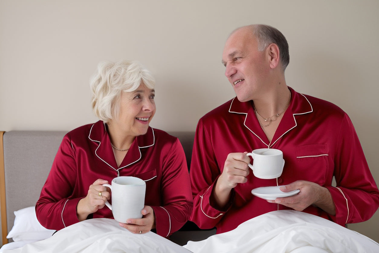 Elderly couple in silk loungewear enjoying coffee in bed, looking blissfully happy.