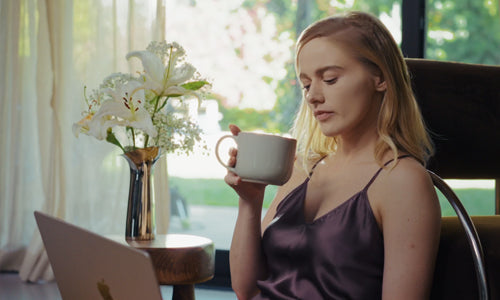 Woman wearing silk sleepwear made for sensitive skin, holding coffee while using a laptop in a calm indoor setting