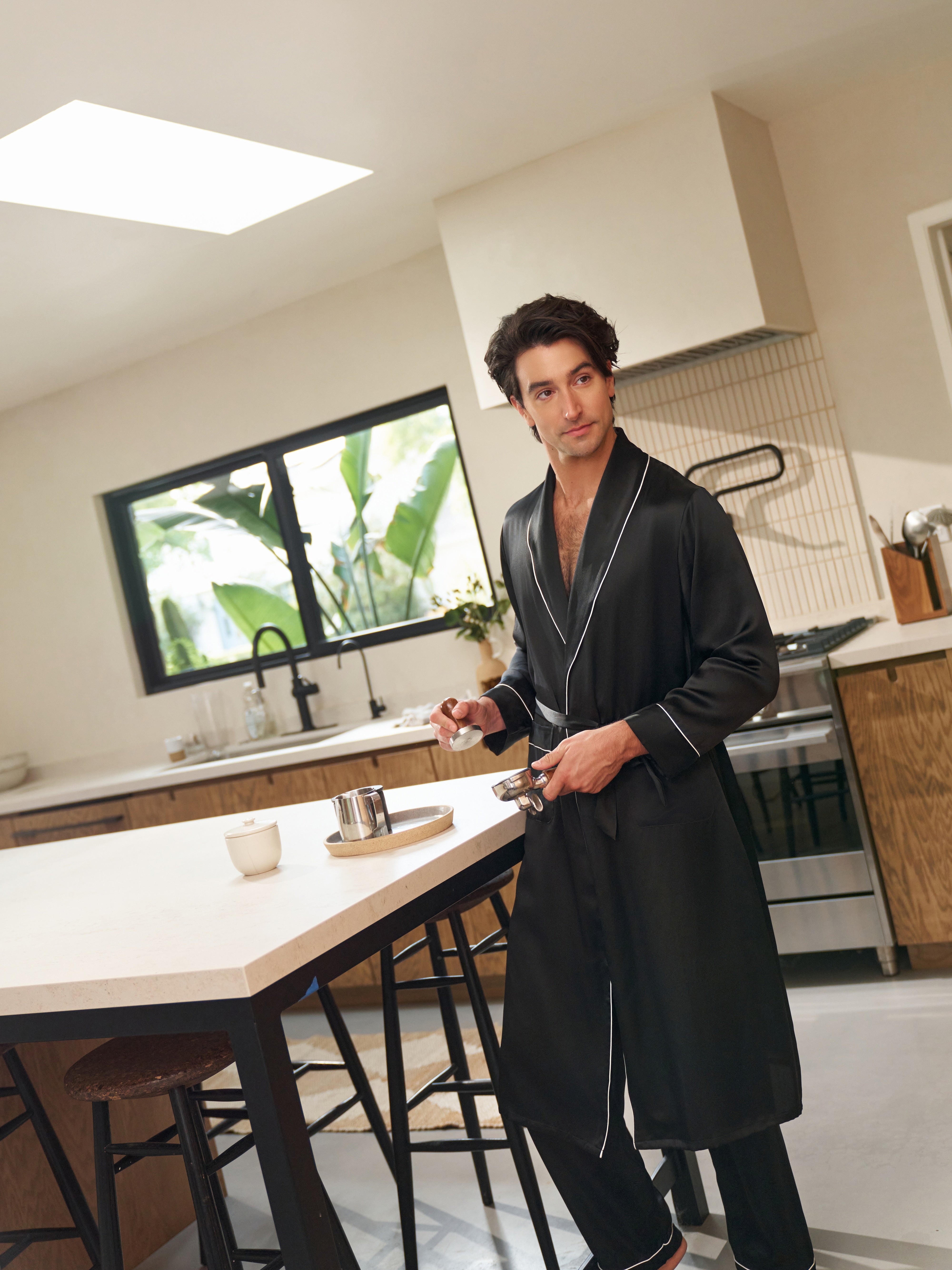 Man wearing Jasmisilk men's black pure silk robe with white piping, preparing coffee in a modern kitchen